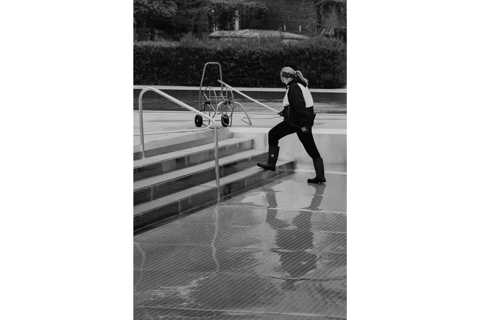Eine Frau steigt aus dem ausgelassenen Schwimmbecken hinaus und hält einen Besen in der Hand. Es is eine Schwarzweißfotografie, Fotoprojekt Strandbad Alte Donau, Wien.