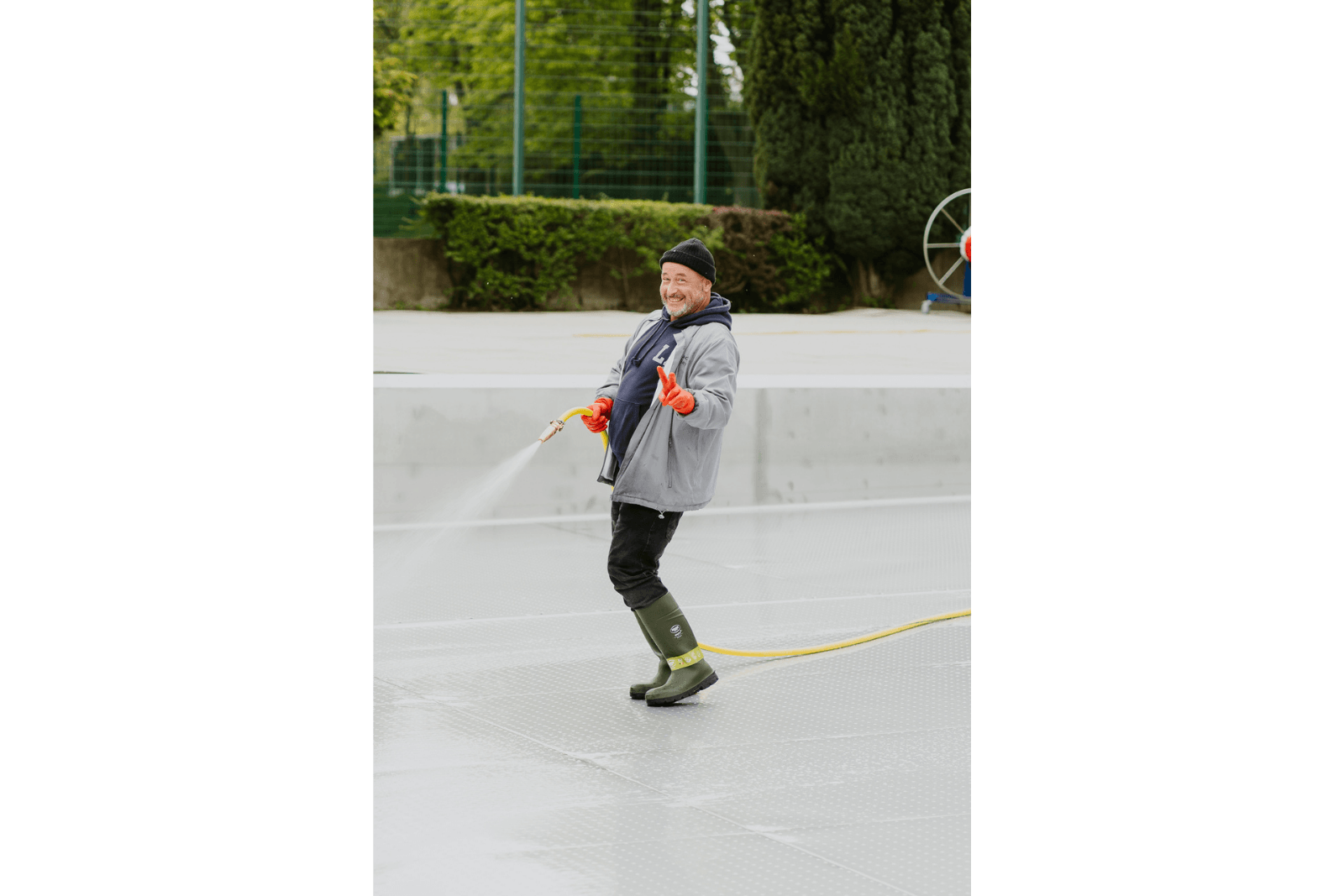 Ein Mitarbeiter reinigt mit Wasserschlauch das Schwimmbecken und grinst fröhlich in die Kamera und formt das Peace Zeichen mit einer Hand.