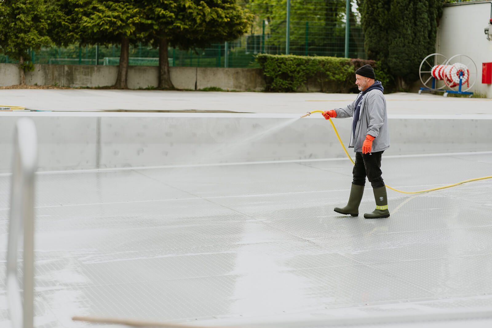 Ein Mitarbeiter reinigt mit Wasserschlauch das Schwimmbecken und grinst fröhlich in die Kamera.