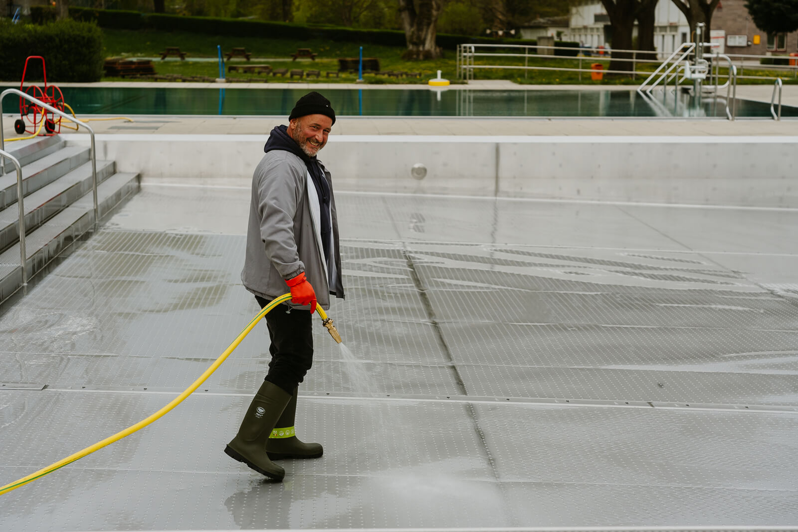 Ein Mitarbeiter reinigt mit Wasserschlauch das Schwimmbecken und grinst fröhlich in die Kamera.