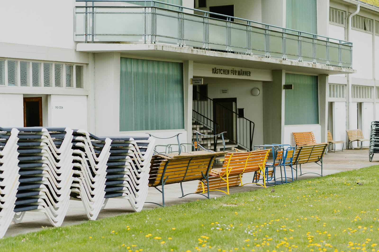 Badehaus Strandbad Alte Donau. Davor stehen Holzbänke und in Stapeln die blauen Liegestühle.