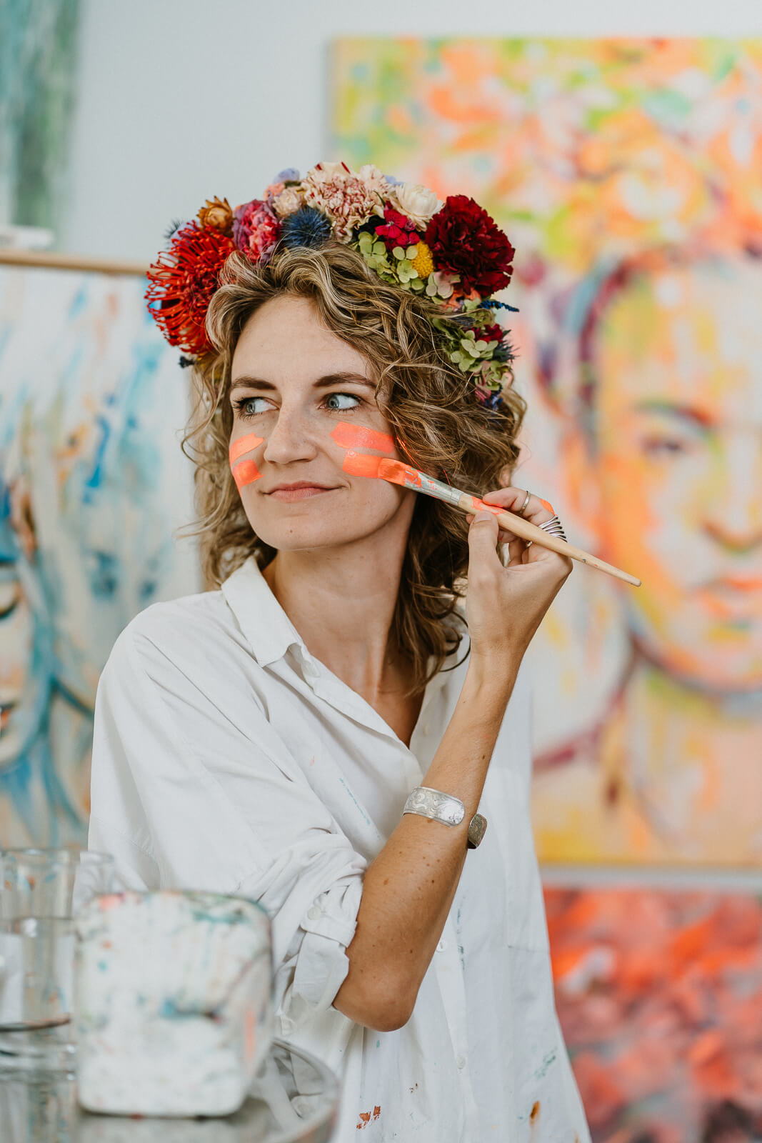 Künstler:innenportrait einer Malerin mit Blumenkranz, mit Pinsel im Gesicht spielend vor ihren Gemälden, kreativ und ausdrucksstark im Atelier inszeniert Select 80 more words to run Humanizer.