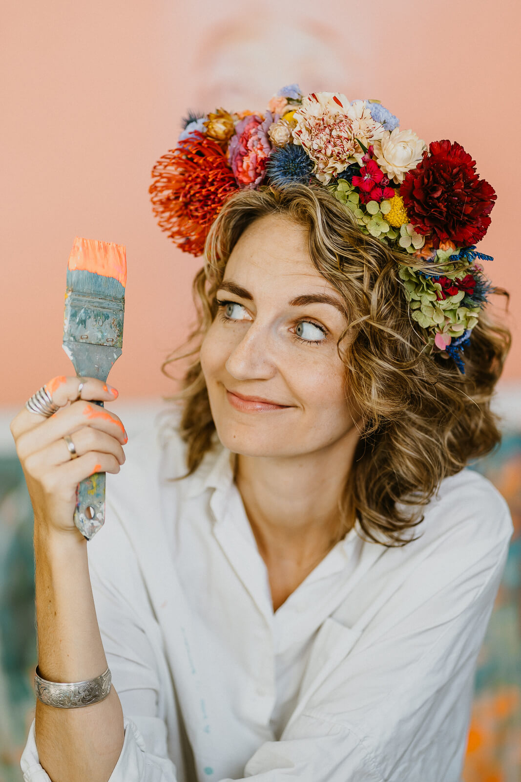 Künstler:innenportrait einer Frau mit buntem Blumenkranz, lächelnd mit einem Pinsel in der Hand, verspielt und kreativ in Szene gesetzt