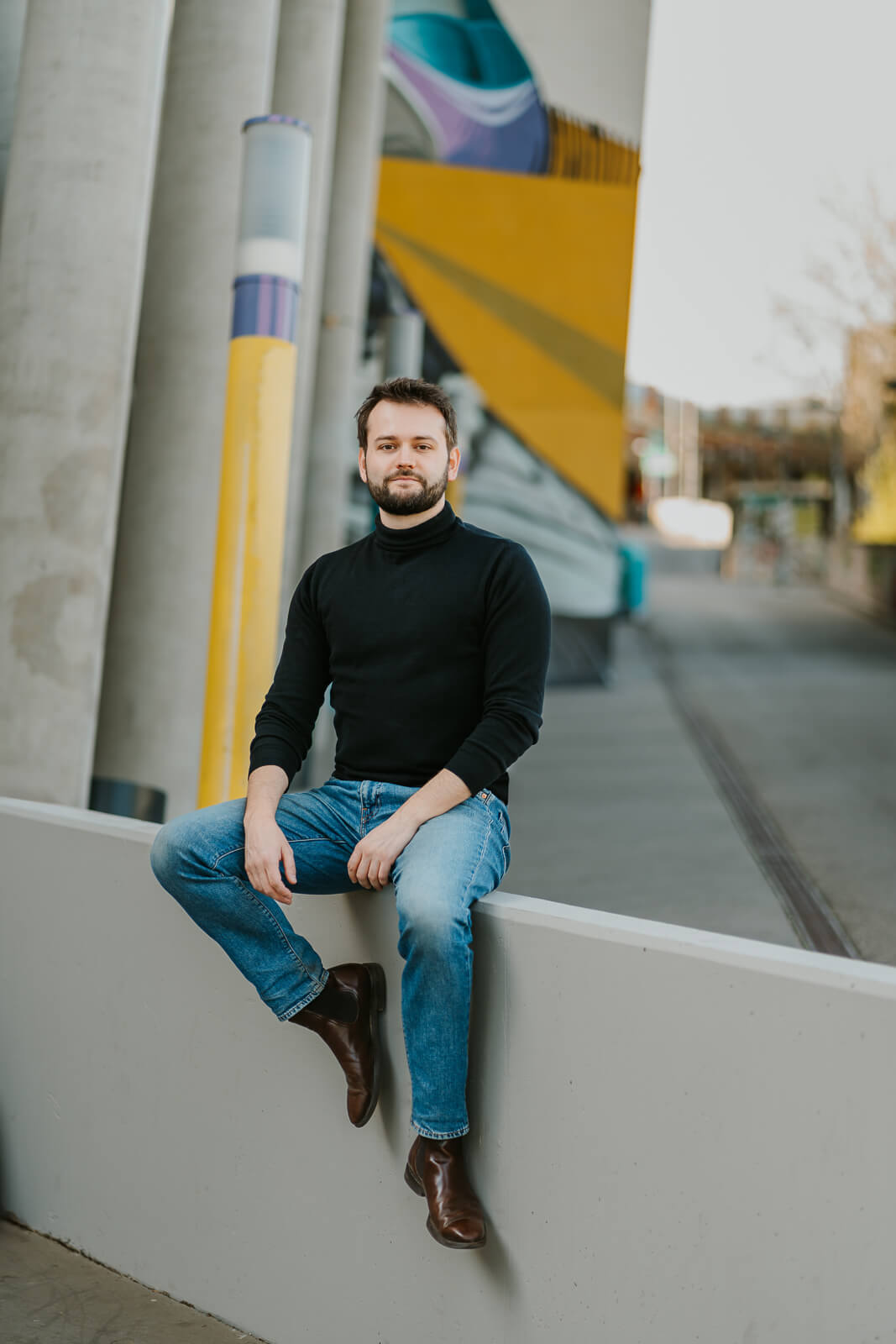 Künstler:innenportrait eines Mannes in urbaner Umgebung, sitzend auf einer Mauer mit ruhigem, selbstbewusstem Ausdruck, natürlich und präsent inszeniert