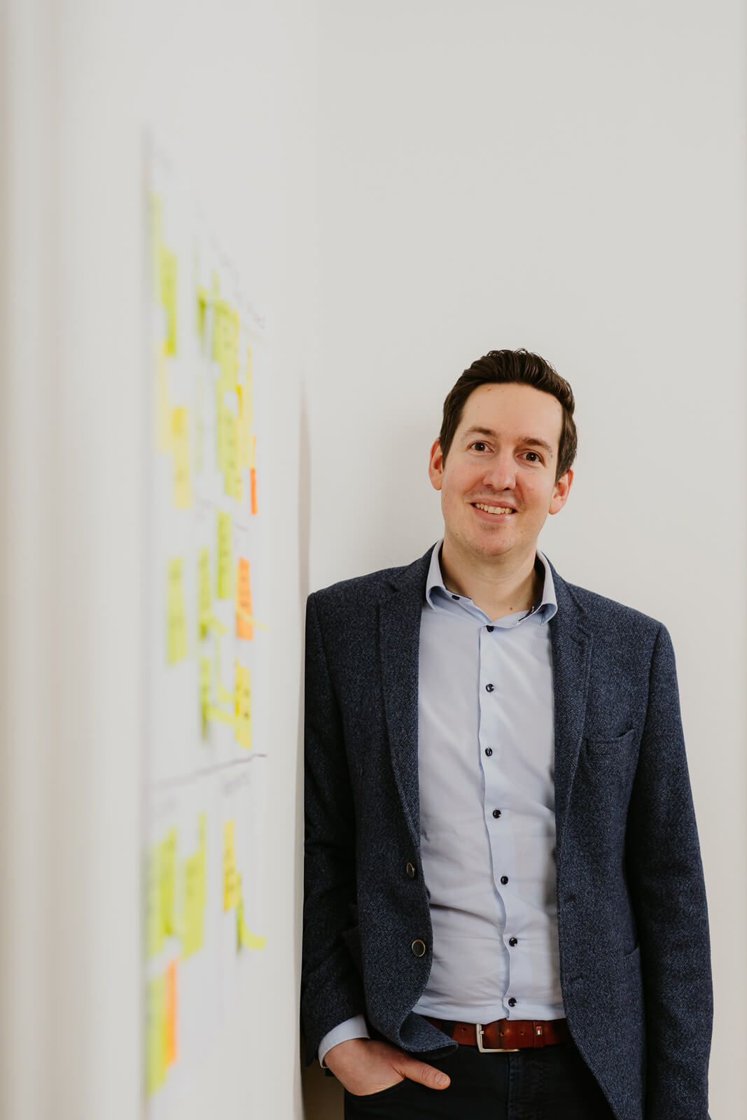 Ein Mann mit blauem Sakko lehnt lässig an der Wand. An der Wand hängt ein Plakat mit Post its. Business und Unternehmensfotografie Wien und Niederösterreich