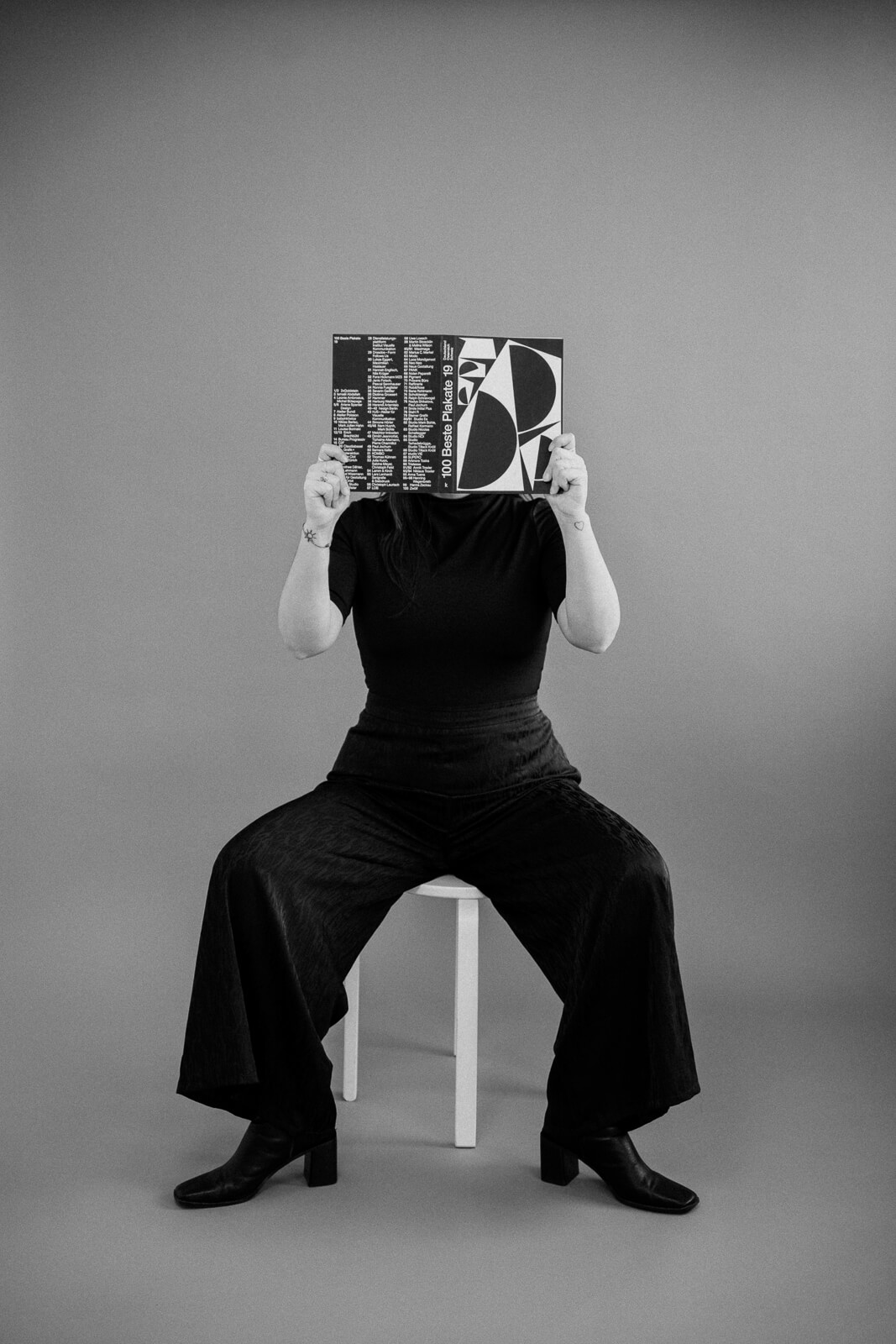 Schwarzweißfotografie einer Frau, die auf einem Sessel sitzt und ein Buch vor ihren Kopf hält.